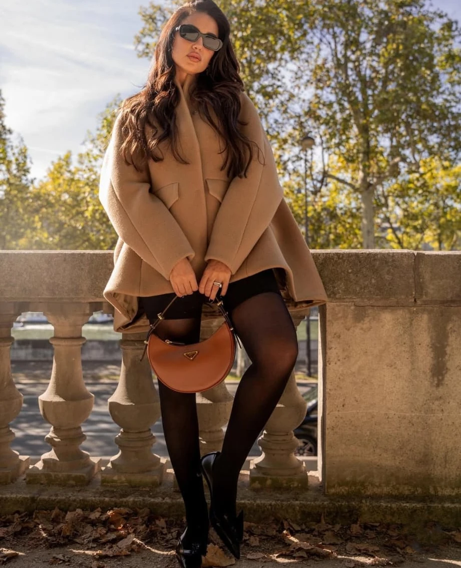 Total look Prada d’une élégance tranquille : cape en laine camel aux lignes sculptées, portée sur mini noire et collants opaques pour un contraste graphique. Escarpins pointus allongent la jambe, tandis que la lunette oversize apporte la note diva contemporaine. Le sac Arqué, en cuir cognac, réchauffe l’ensemble et dialogue avec la lumière dorée de l’automne. Silhouette équilibrée, chic parisien assumé, parfaite du matin au rendez-vous pro. Astuce style : relever le col et jouer sur des gants fins pour accentuer la dimension couture. Maquillage doux, cheveux libres impeccables également. dora-zarouk-prada